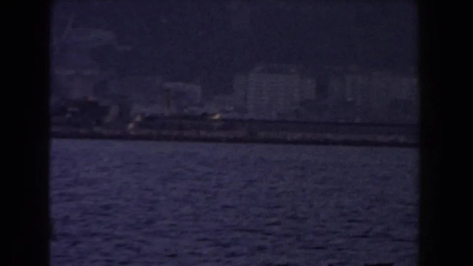 Distant Gibraltar shoreline and waterfront structures seen across blue water in grainy 1959 8mm footage.