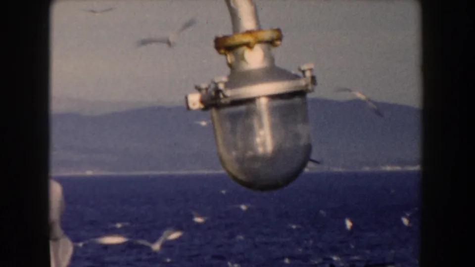Boat-mounted lamp fixture in foreground as white birds fly over the blue sea near Gibraltar.