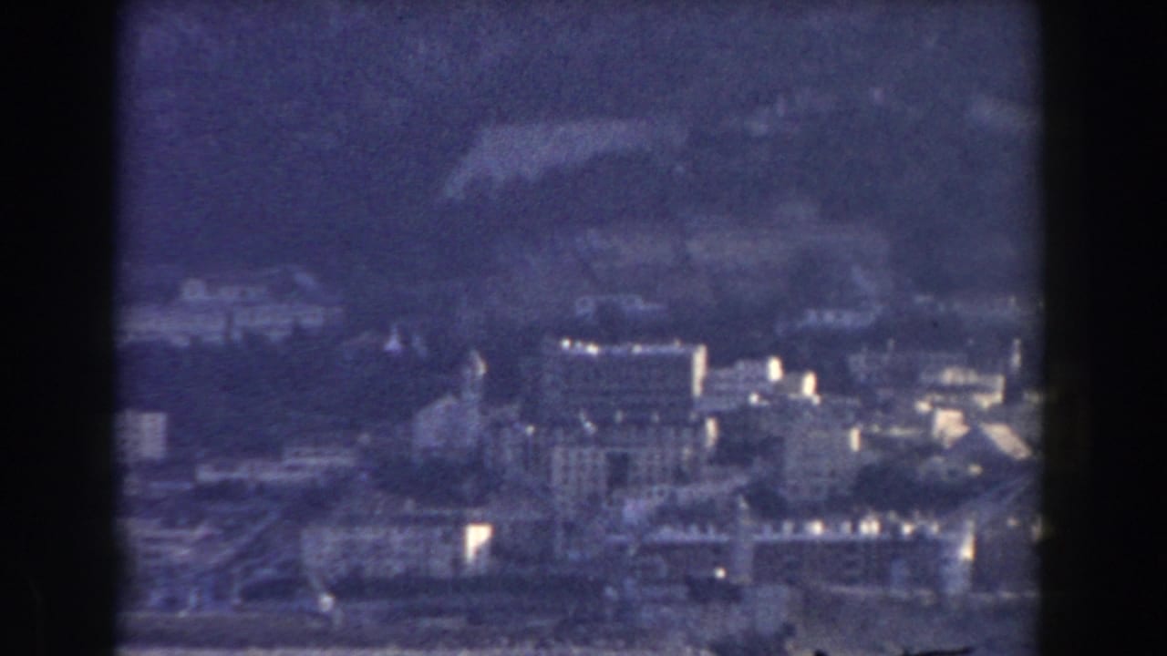 Distant city buildings below a dark mountain slope, seen through a soft blue 8mm frame.