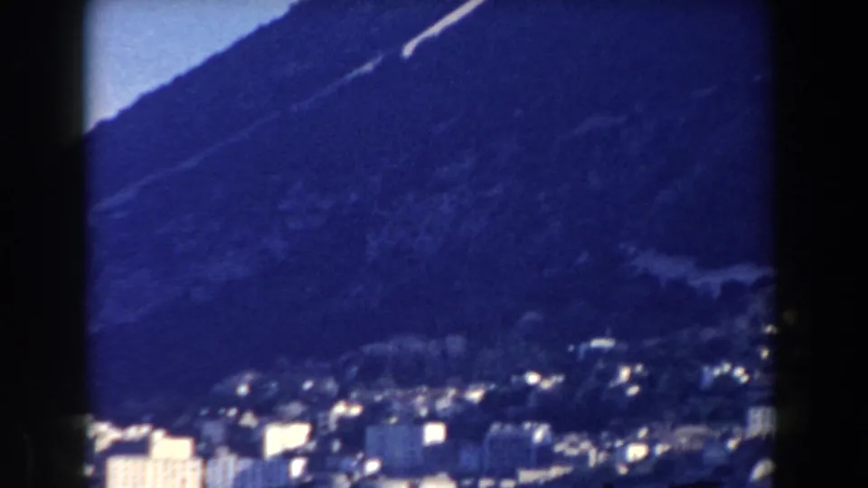 Late pan frame from 1959 Gibraltar with small bright buildings below a steep blue-toned mountain slope.