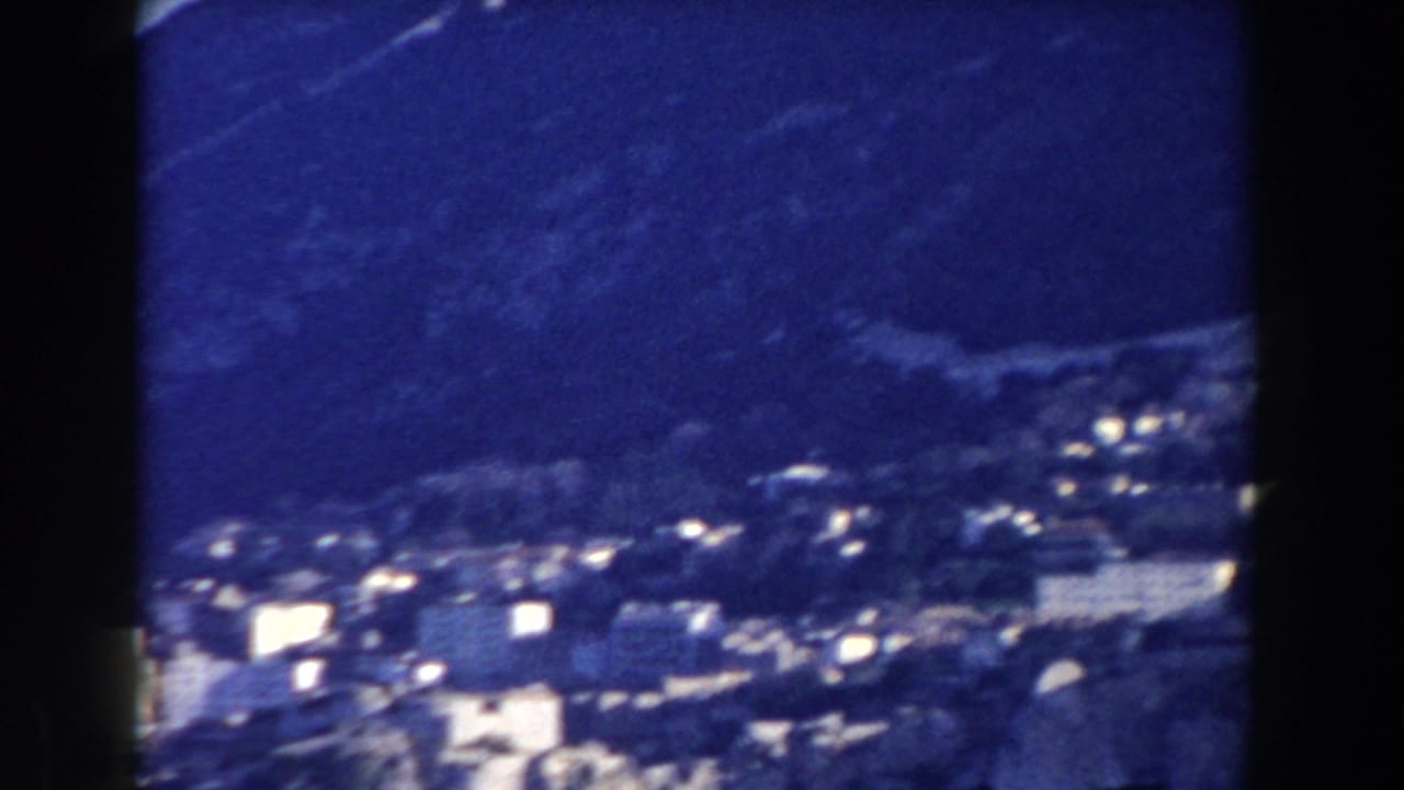 Distant hillside town below dark mountain slopes on blue-shifted 8mm film.
