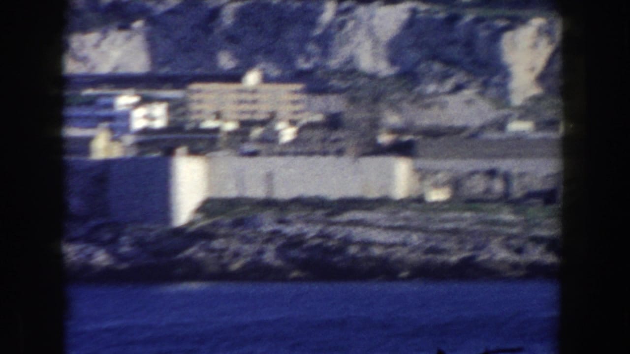 White coastal buildings below rocky cliffs with blue water in the foreground.