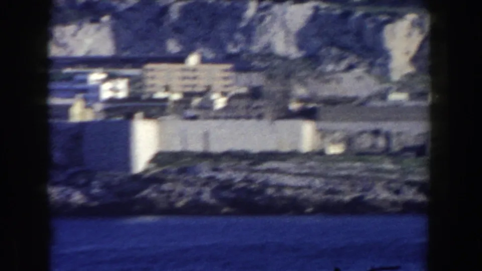 1959 Gibraltar coastal view with white buildings, rocky shoreline, blue water, and pale cliffs seen through an 8mm frame.