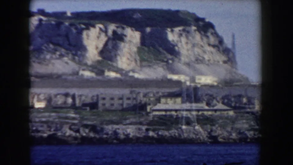 Gibraltar shoreline in 1959, showing low coastal buildings, rocky ground, blue water, and a high limestone cliff in soft 8mm color.