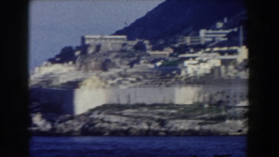 1959 Gibraltar coastal buildings and pale waterfront walls appear below a shadowed hillside, with water across the foreground.