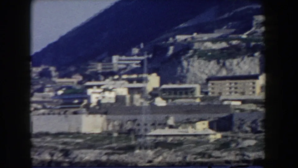 Gibraltar waterfront in 1959 with white buildings, retaining walls, rocky terrain, and a steep hillside captured on 8mm film.