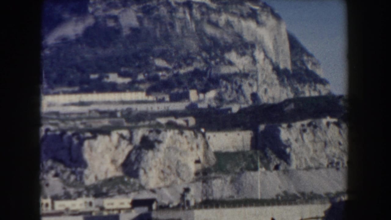1959 Gibraltar home-movie frame showing the Rock's limestone slopes, coastal walls, and dark blue water in the foreground.