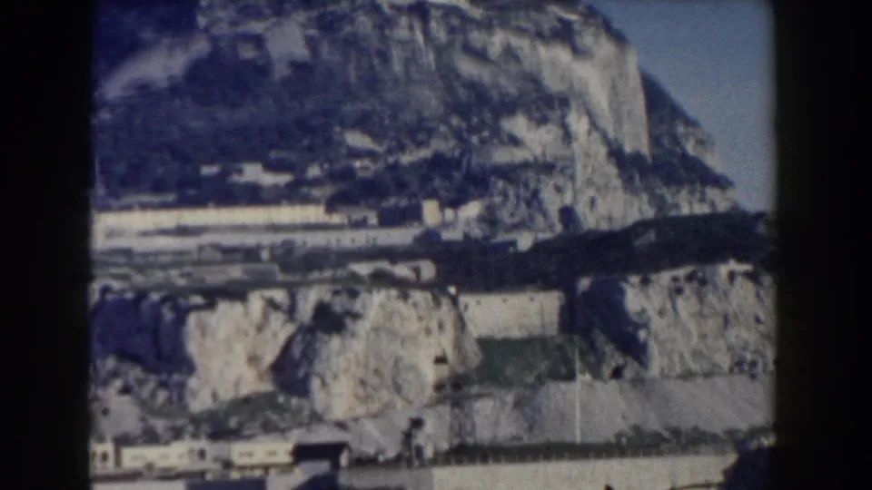 1959 Gibraltar home-movie frame showing the Rock's limestone slopes, coastal walls, and dark blue water in the foreground.