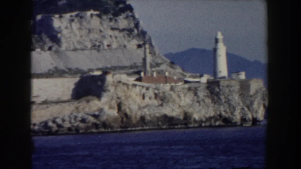 Gibraltar shoreline view from 1959 showing a white tower, stone walls, rocky headland, and distant hills beyond the water.