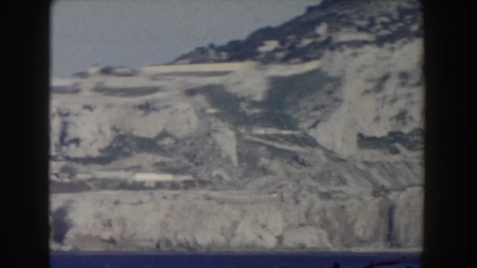 Soft 1959 Gibraltar home-movie frame showing pale limestone slopes, waterfront structures, and the sea from a distance.