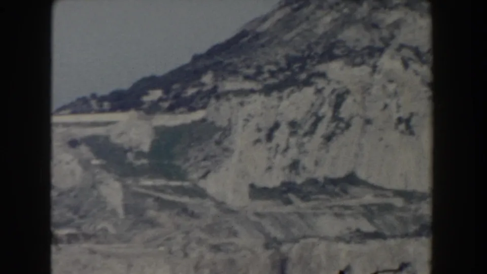 1959 Gibraltar 8mm frame showing pale rock faces, dark seams, and a steep mountain slope under a washed sky.