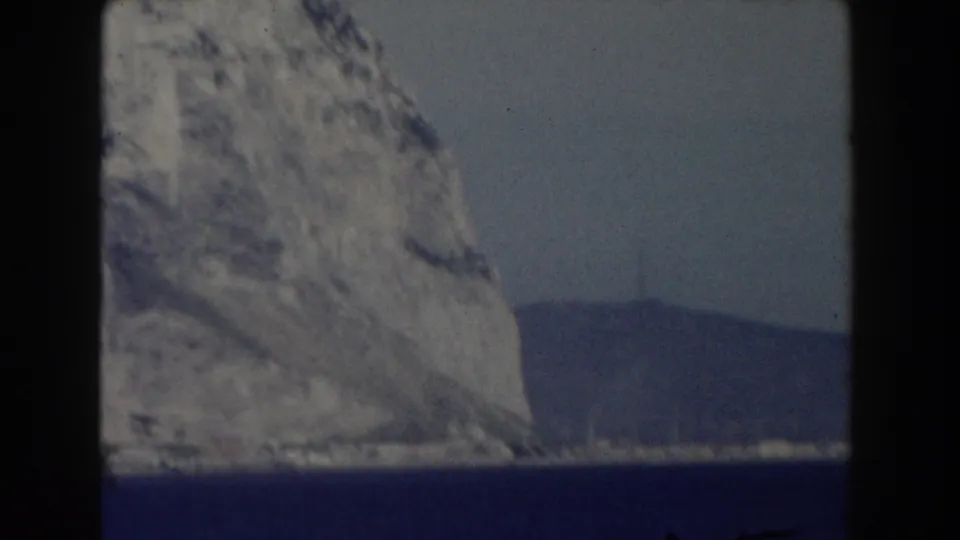 Gibraltar 1959 home-movie frame with a cliff edge at left, deep blue water below, and distant land across the sea.