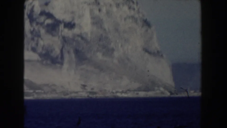 1959 Gibraltar frame showing a broad limestone cliff rising from dark blue sea, with a distant shoreline visible to the right.