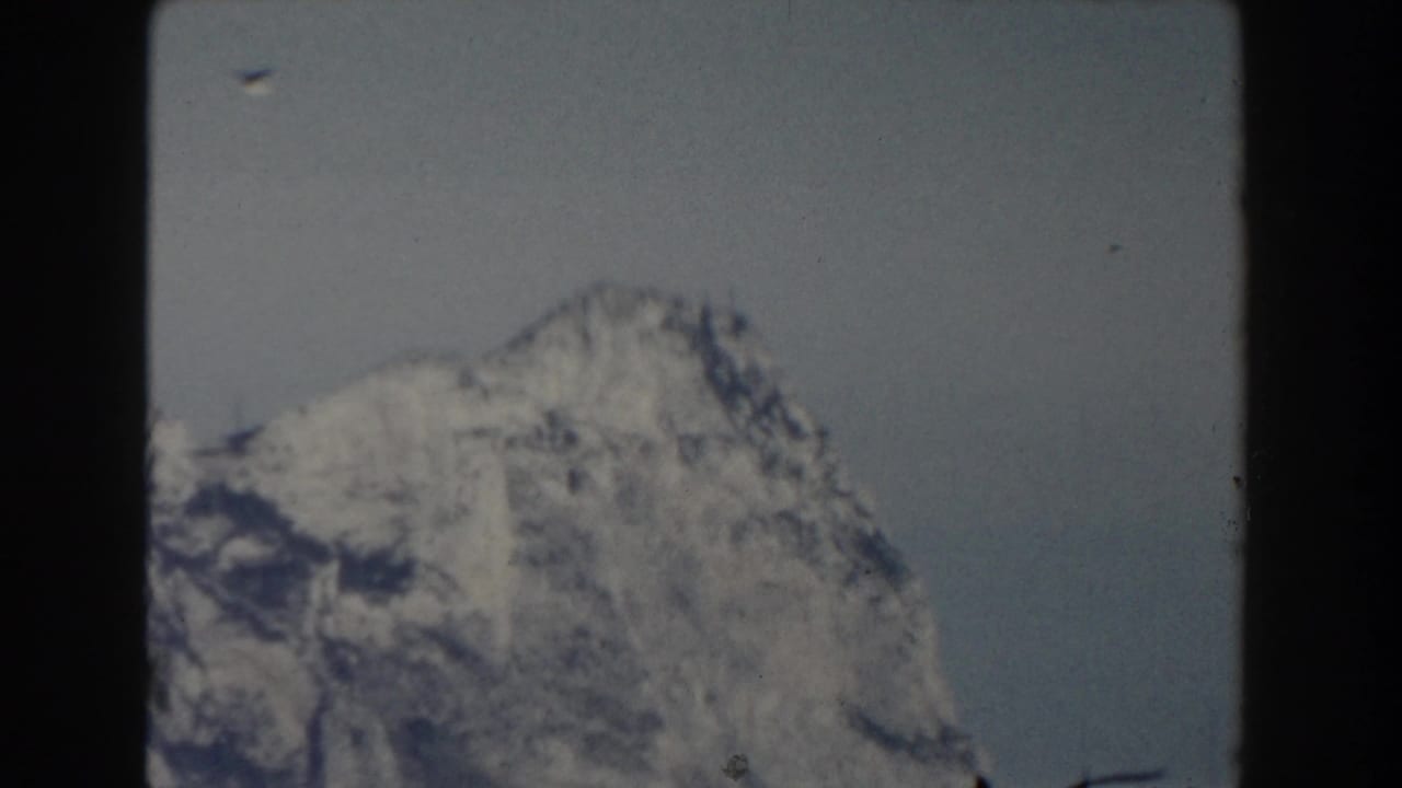 1959 8mm frame of a rugged snow-covered mountain peak, softly exposed against a muted sky.