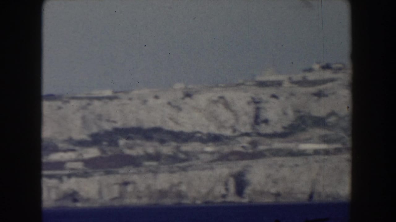 1959 8mm frame showing the Rock of Gibraltar from across the water, with pale cliffs and scattered shoreline buildings.