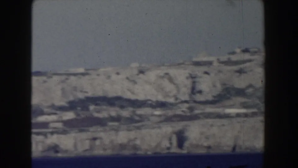 1959 8mm frame showing the Rock of Gibraltar from across the water, with pale cliffs and scattered shoreline buildings.
