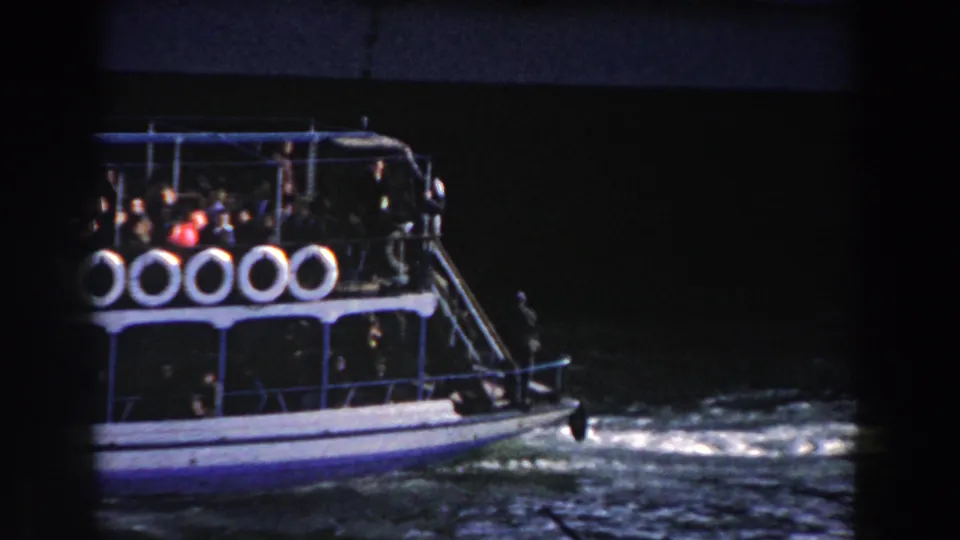 The forward section of the blue and white ferry moves through dark harbor water, with passengers gathered behind the rail.