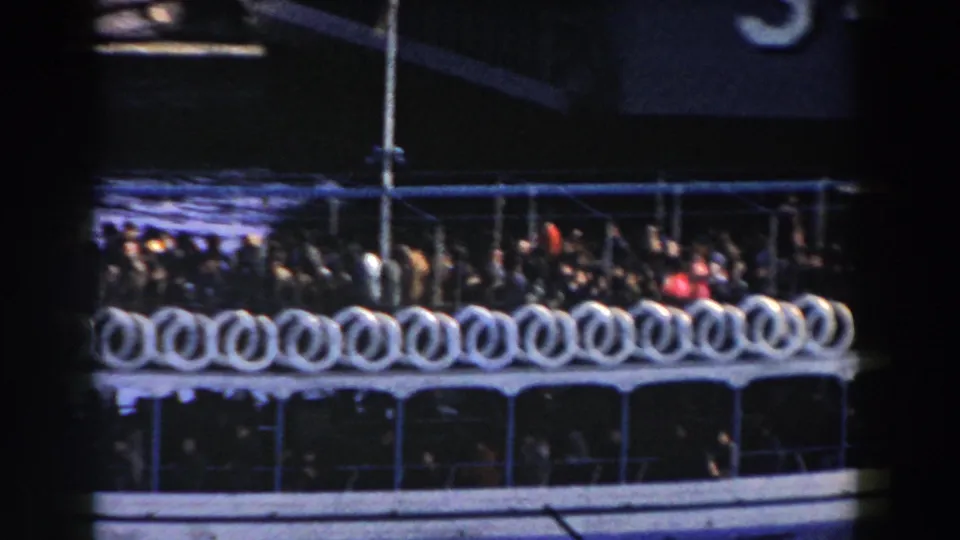 A crowded blue and white ferry continues past a dark ship structure, its rail lined with many white life rings.