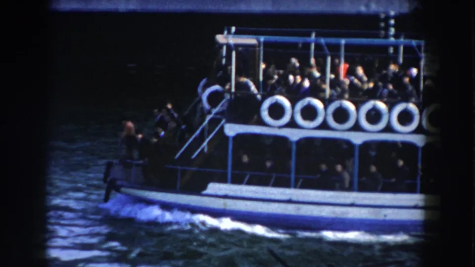 1959 Gibraltar ferry footage showing the vessel's side and stern area, with wake below and passengers seated or standing on two decks.