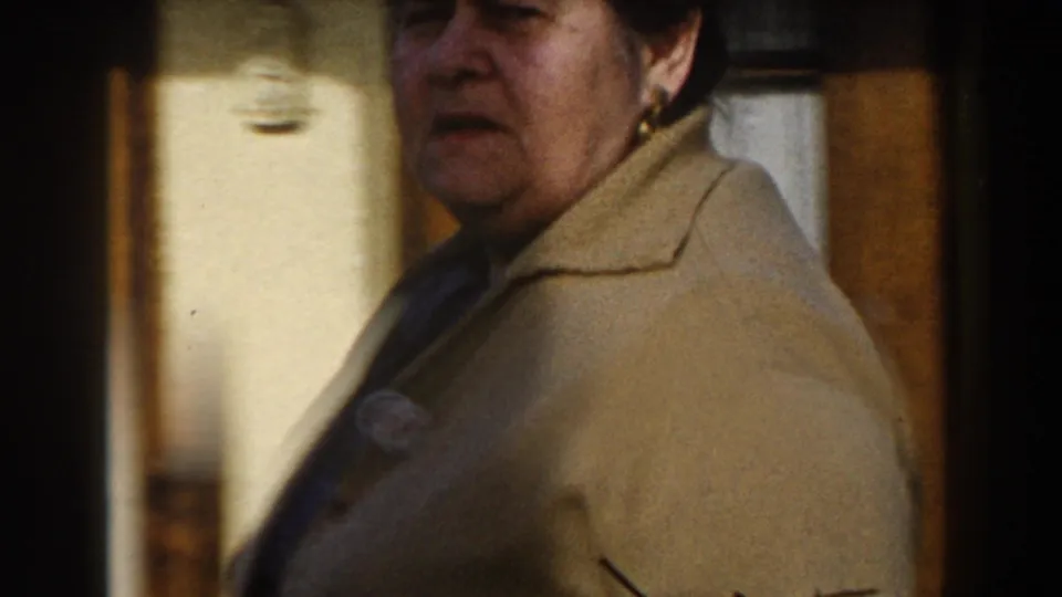 Brief 1959 travel-film close-up of a woman in a beige coat, framed softly near a doorway or interior wall.