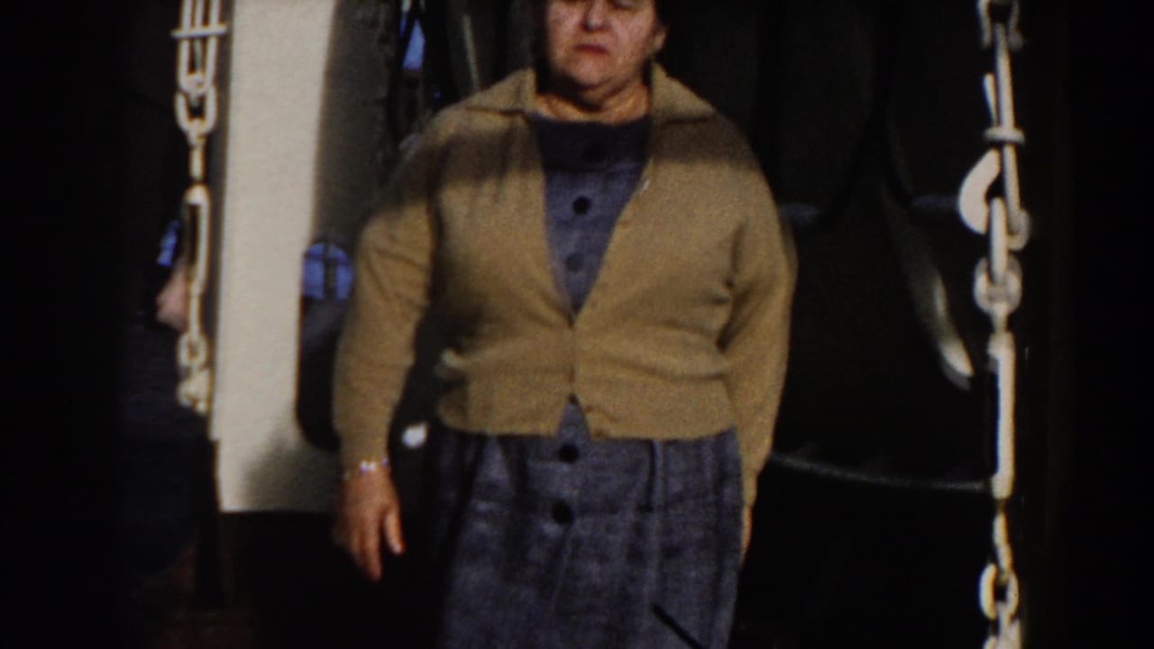 Older woman stands on a ship deck between chains and dark equipment.