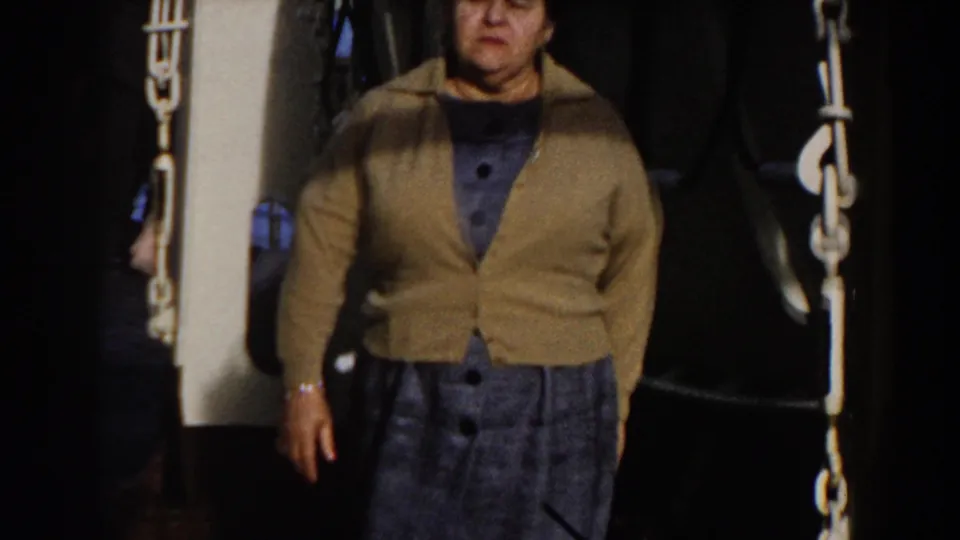 Older woman stands facing the camera on a ship deck, with heavy chains and dark industrial fittings around her.