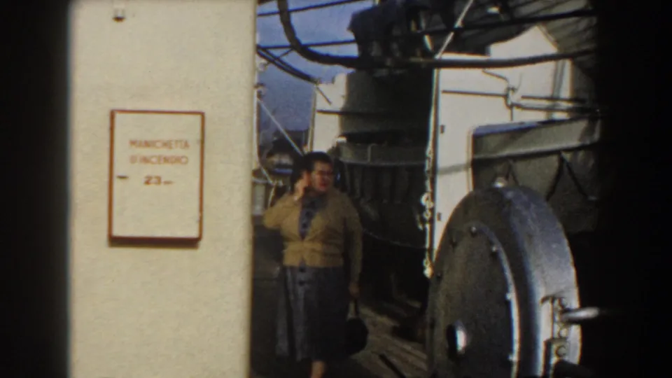 Wider ship-deck view with the woman beside large equipment, white chains, and a readable Italian fire-hose sign.