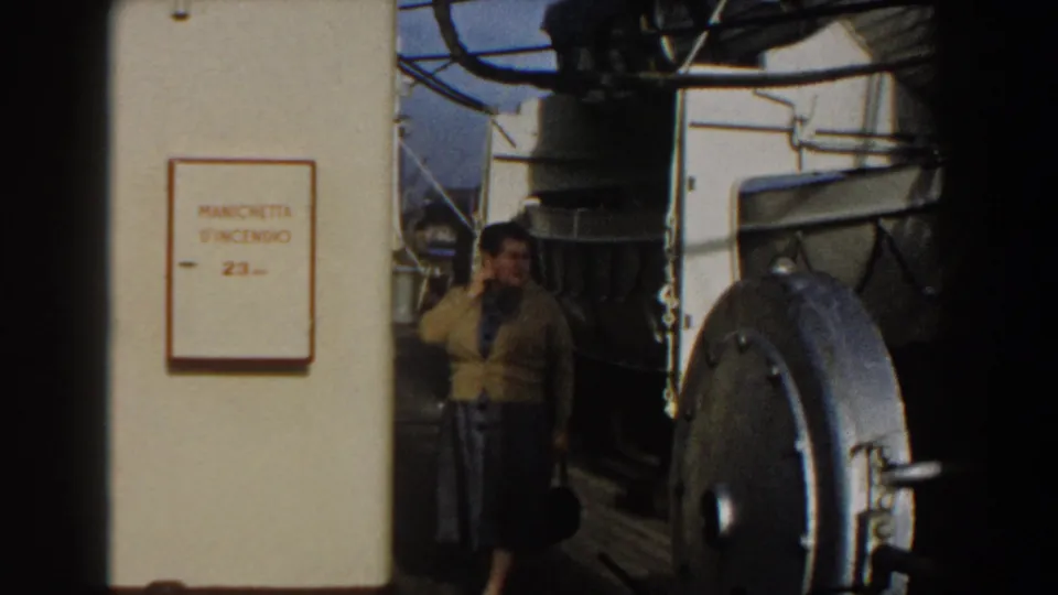 In Gibraltar 1959, a woman in a yellow sweater stands on a ship deck near an Italian fire-hose cabinet sign and metal hatch.