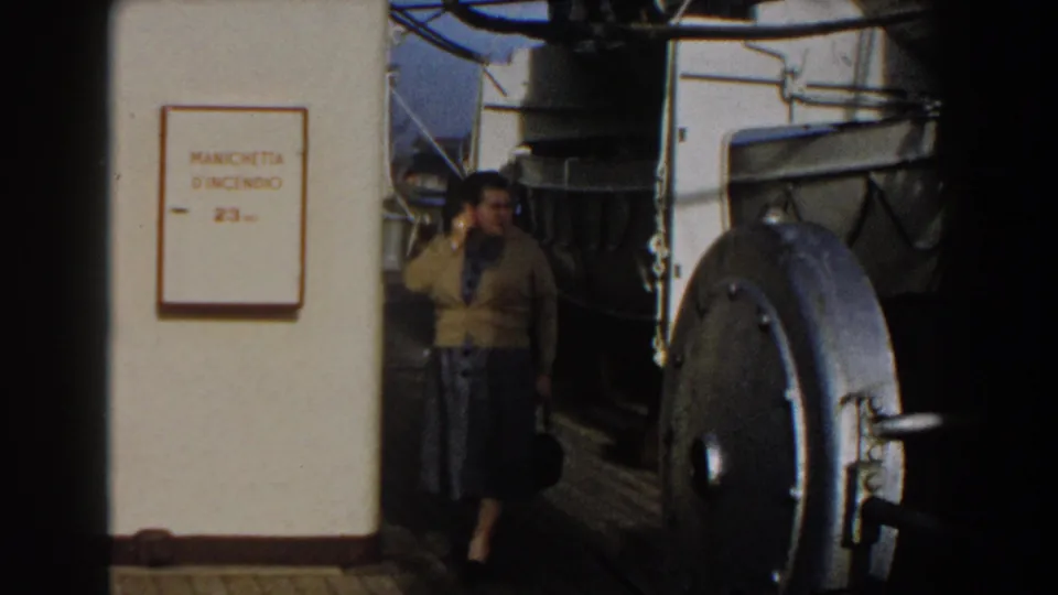 A woman in a yellow cardigan and dark skirt walks along the 1959 Gibraltar ship deck beside the fire-hose cabinet sign.