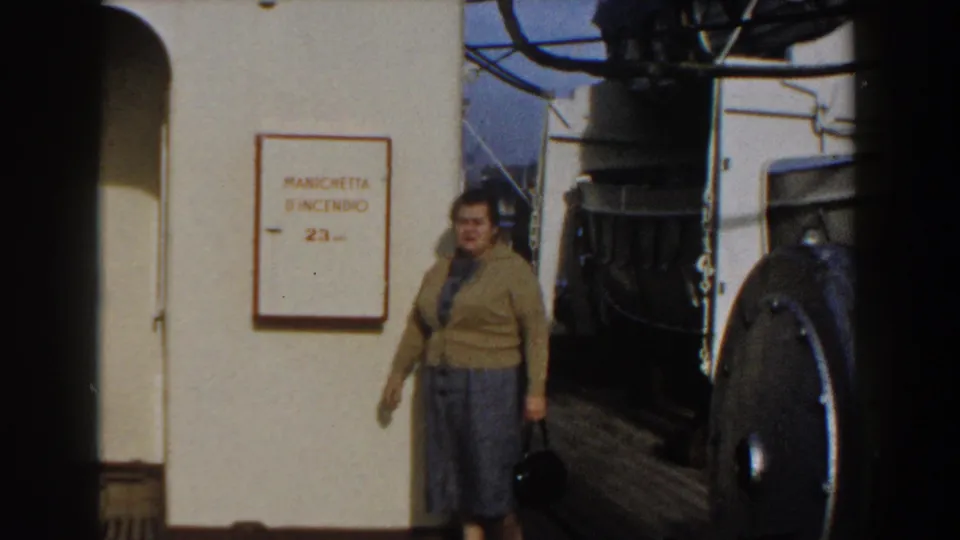 The woman carries a black bag across the Gibraltar ship deck, with the fire-hose sign and large metal fitting behind her.