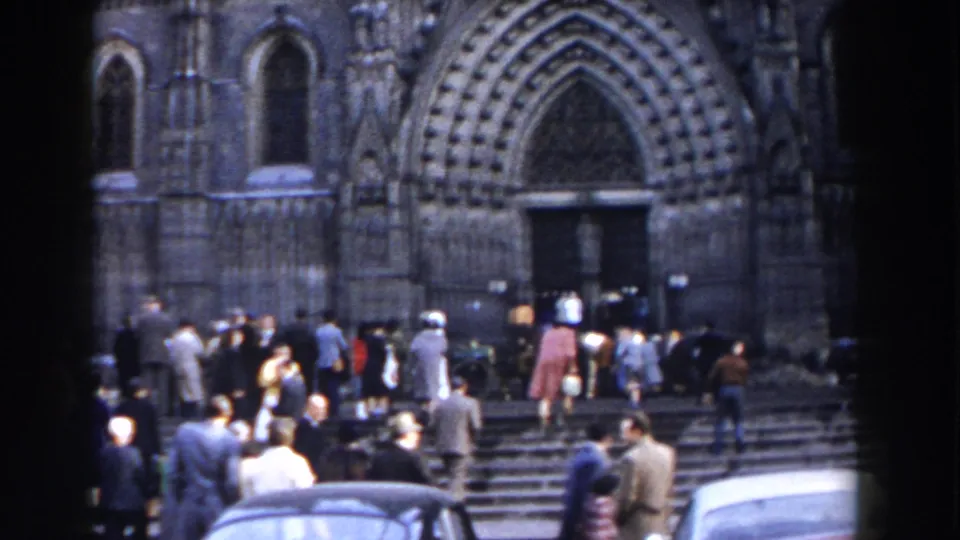 A crowd disperses or gathers at the steps of a large Gothic-style church in 1959 Gibraltar footage, with cars partly visible below.