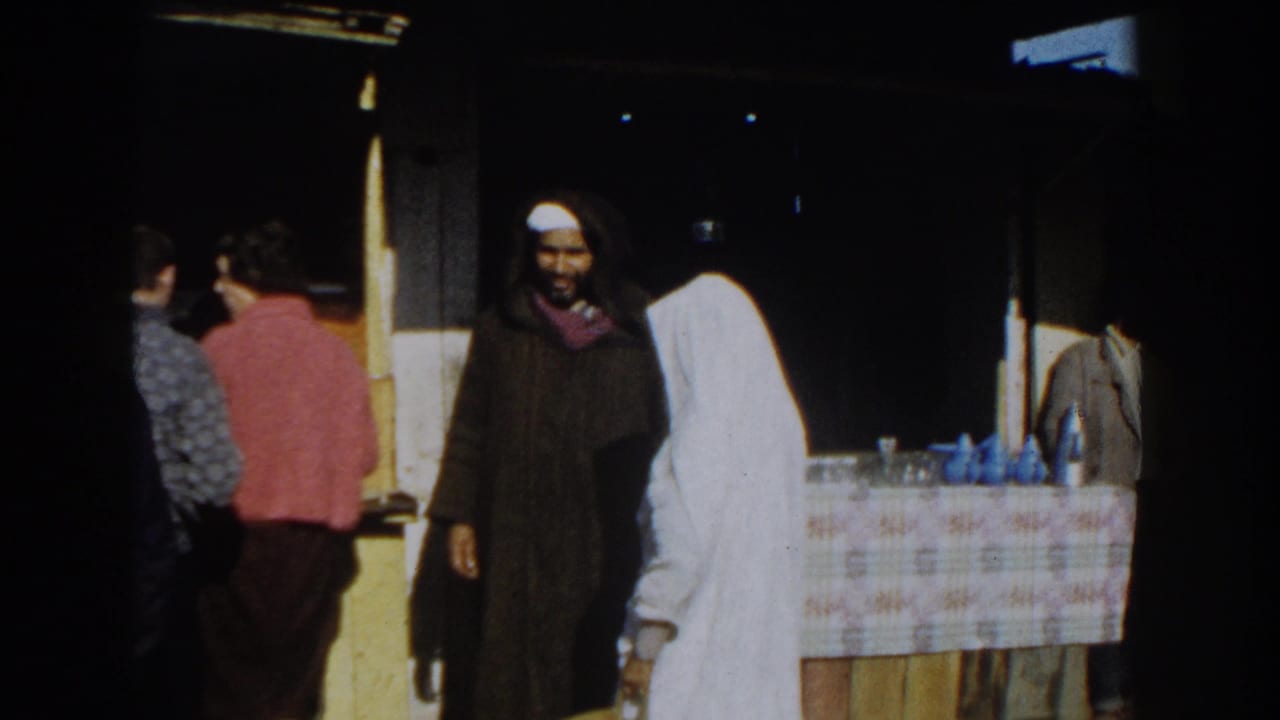 People stand and walk near an outdoor market stall with a checkered table covering.