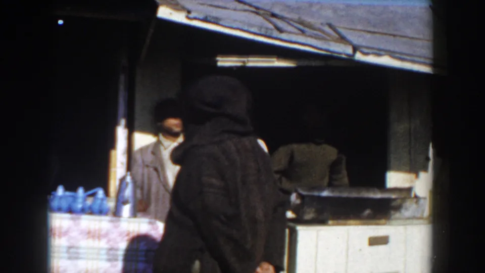 1959 Casablanca home-movie frame showing men near a rustic stall, a hooded figure in front, and blue jugs on a patterned counter.