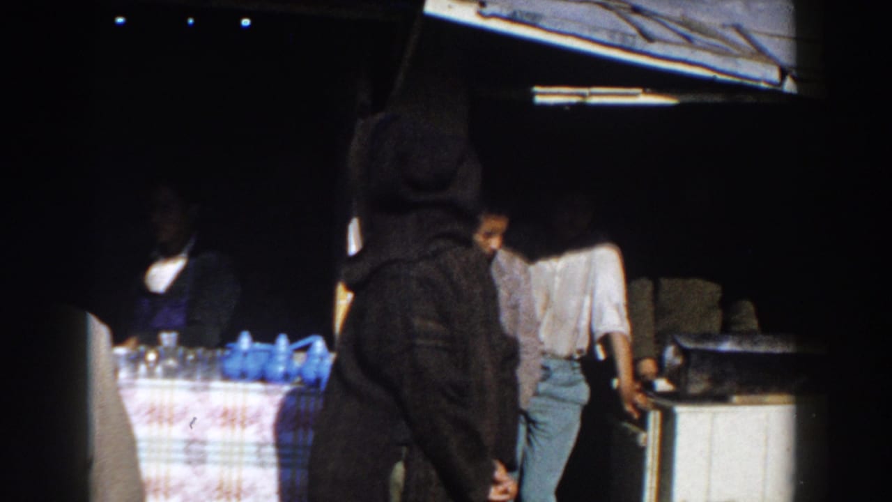 A hooded figure stands before a stall while men in shirts gather beside a counter.