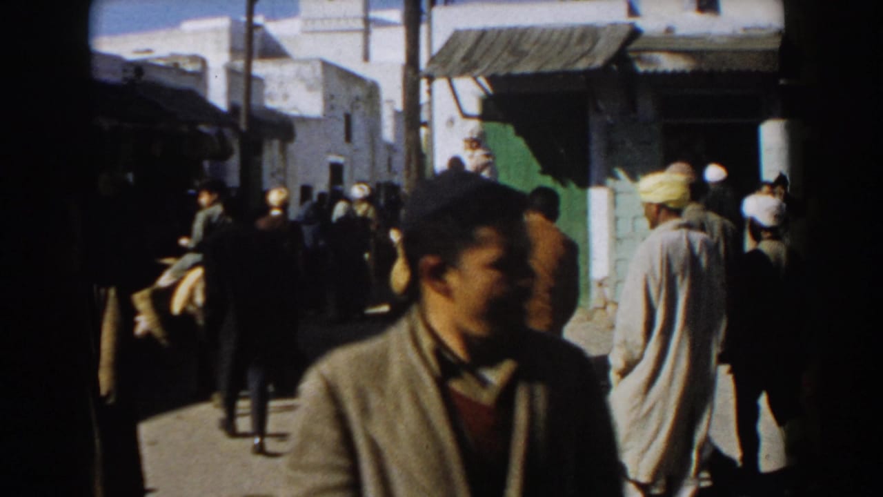 Men walk along a busy narrow street with a donkey rider and white buildings in the background.