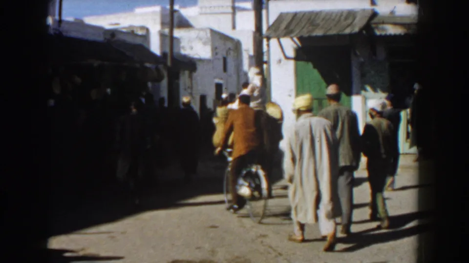 A handheld 8mm view down a Casablanca street shows pedestrians in robes and coats walking away, with a bicycle rider near shaded storefronts and white walls.