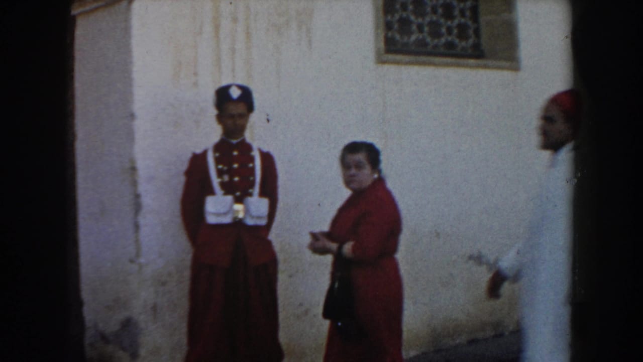 Guard in red uniform stands beside a woman near a weathered wall and decorative window.