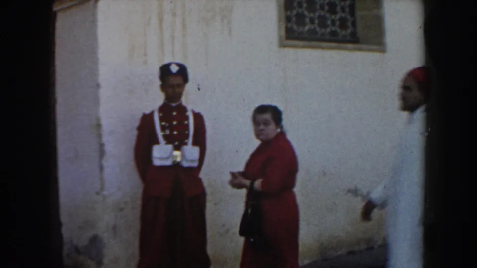In metadata-supported 1959 Casablanca, a red-uniformed guard stands near a woman in red as passersby move along a wall with a decorative grate.