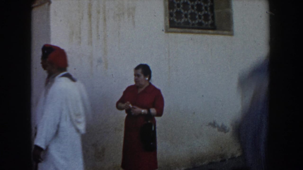 Man in white robe and red cap walks past a woman in red near a wall.