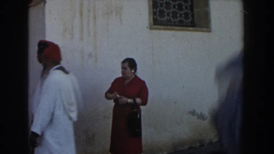 A man in a white robe and red cap passes a woman in red near a weathered wall and decorative window grate in Casablanca home-movie footage.