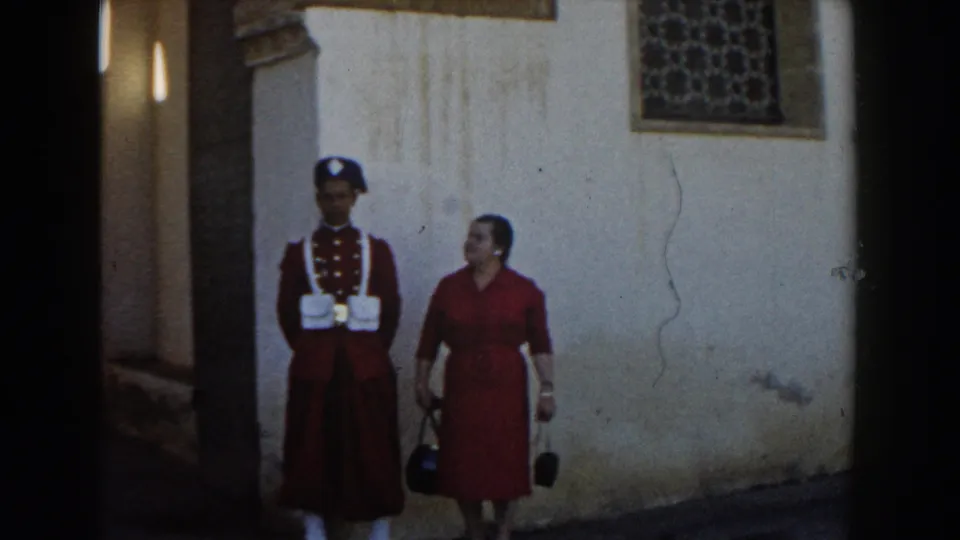 The guard and woman stand side by side for the camera near a pale wall and decorative window, with narrow passage shadows at the left edge.