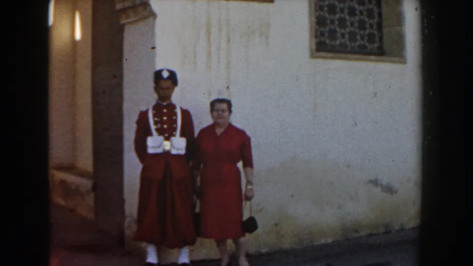 A guard in red uniform and a woman in a red dress pose close together against a weathered wall in metadata-supported 1959 Casablanca footage.