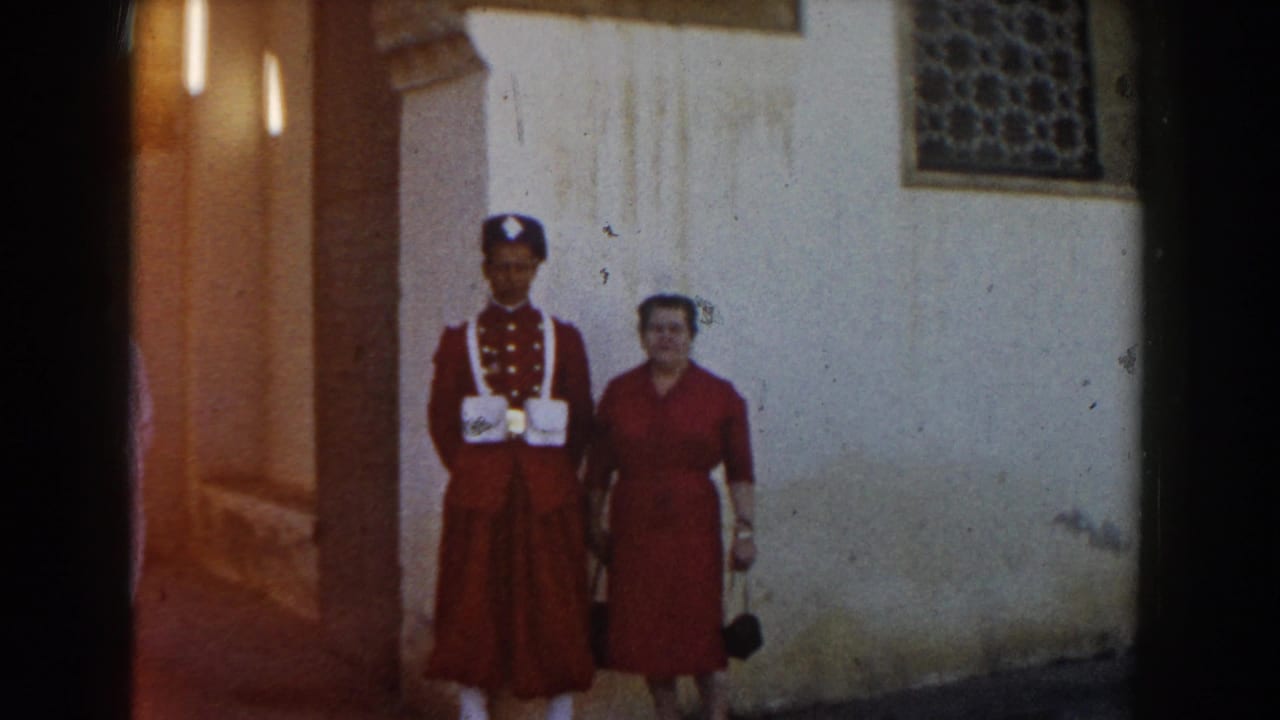 In Casablanca 1959 home-movie footage, a woman in red stands beside a uniformed man in red and white near a stucco wall and patterned window.
