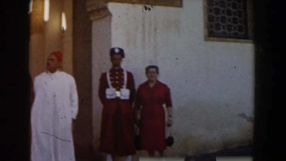 A man in a white robe and red cap walks across the foreground as the woman and uniformed man continue posing by the Casablanca wall.