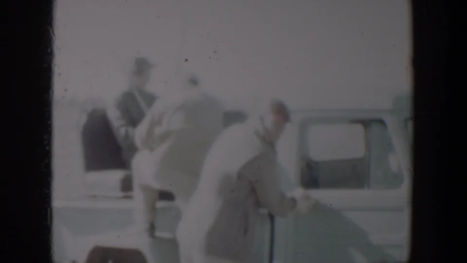 A person bends beside the pale pickup as other adults stand near the truck bed in the supplied Lubbock 1957 clip.