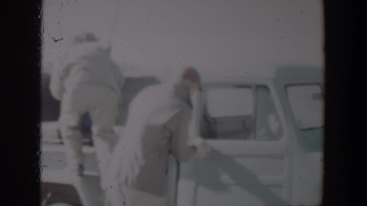 Two men in coats stand beside a light-colored pickup while a rifle is handled near the cab.