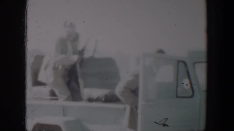 In the pickup bed, an adult man appears to hold a long rifle while another figure bends near the cab, consistent with hunting preparation.