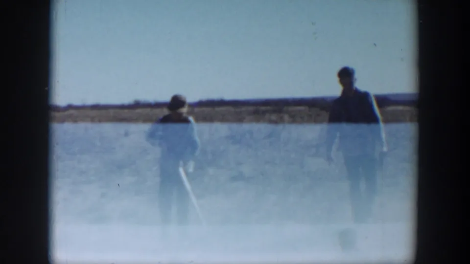 Two distant figures appear in silhouette against a bright sky and low horizon, partially veiled by exposure flare in the 8mm frame.