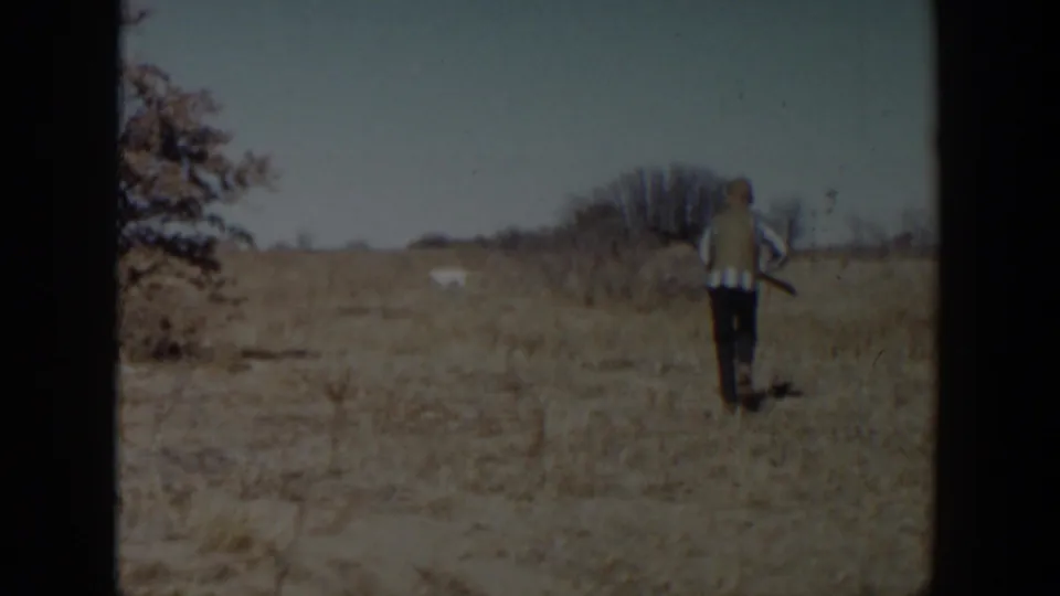 1957 Lubbock-area 8mm frame showing dry grass, trees, and a small distant upright figure or object in the field.