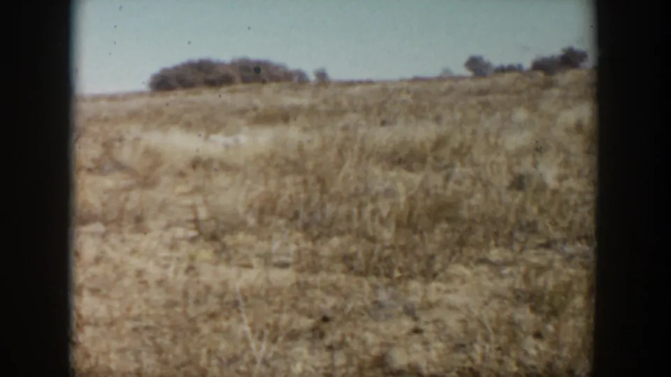 1957 Lubbock, Texas 8mm frame showing dry prairie grass, pale soil, distant shrubs, and clear sky from a moving viewpoint.